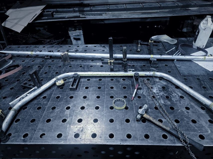 A long bent tube lies on the fixture table beside a tape measure and a hammer