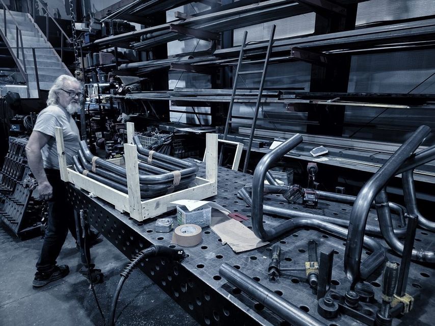 A man with white hair lifts a wooden crate frame beside bent tube parts on the table