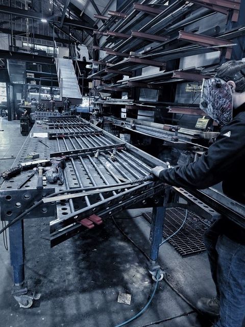 A welder in a hood works on a railing panel of round bar on the fixture table
