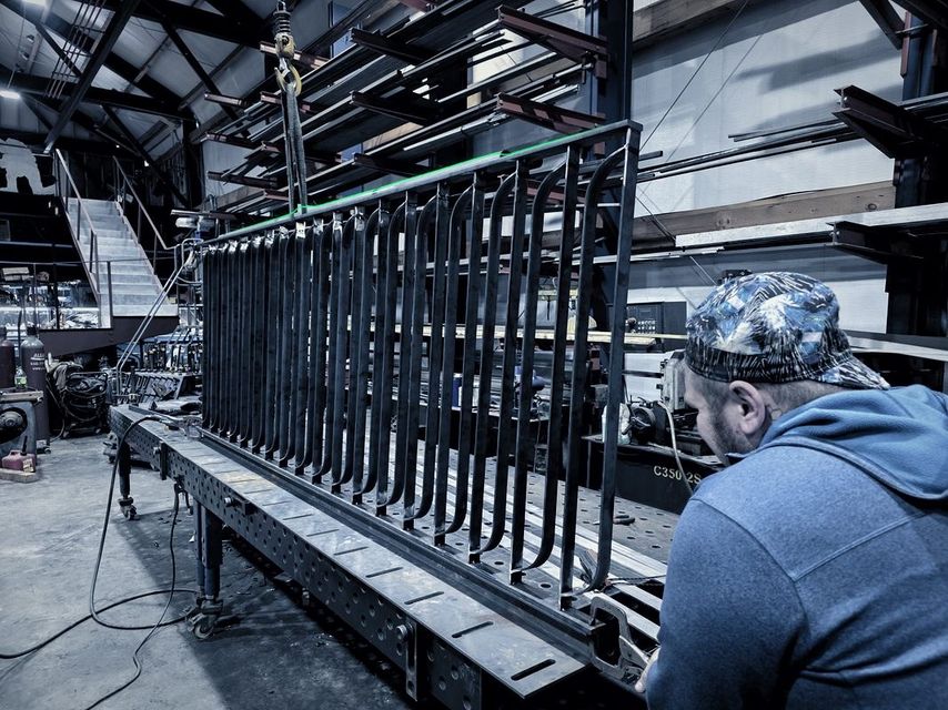 A man works on a steel railing panel with curved pickets standing on the fixture table